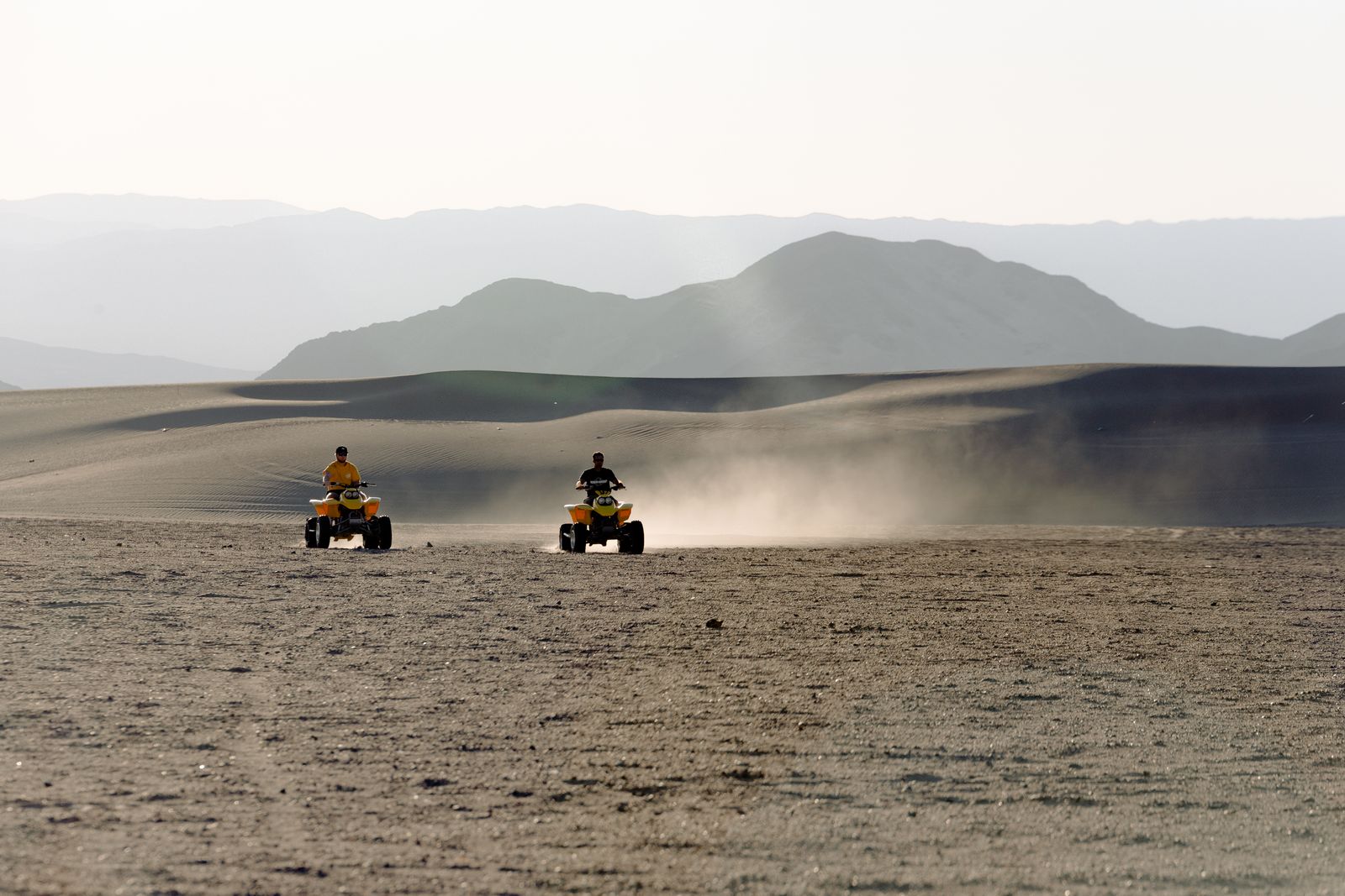 Two riders on ATVs crossing desert sand with mountains behind